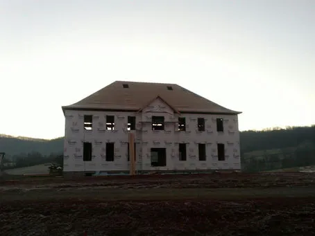A partially constructed house with exposed framing, wrapped in white protective sheeting, set against a dawn sky