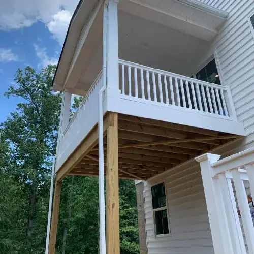 A white, two-story house with a wooden deck supported by pillars, surrounded by trees and a blue sky