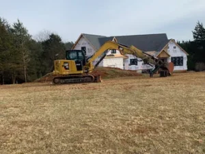 A yellow CAT excavator sits on a dirt mound in a field, with a partially built house wrapped in white house wrap