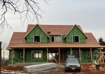 Front view of a large two-story house under construction on a muddy lot, wrapped in green house wrap