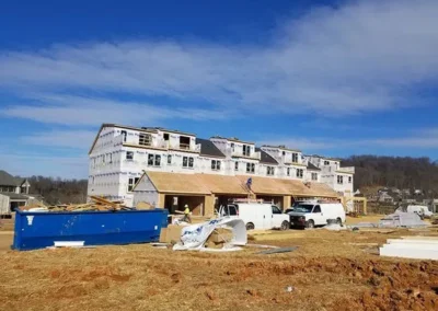 Row of townhouses under construction, wrapped in Tyvek, with exposed framing on the ground floor, construction vans, and a large blue dumpster