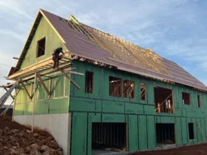 Two-story house under construction with a steep gable roof, wrapped in green sheathing, as a worker installs roofing material