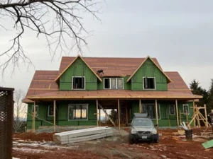 a large two-story house under construction on a muddy lot, wrapped in green house wrap with the roof